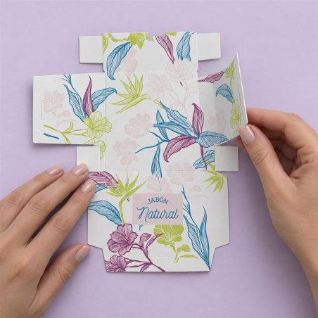 Manos plegando las solapas de una caja para jabón con estampado floral sobre fondo lila